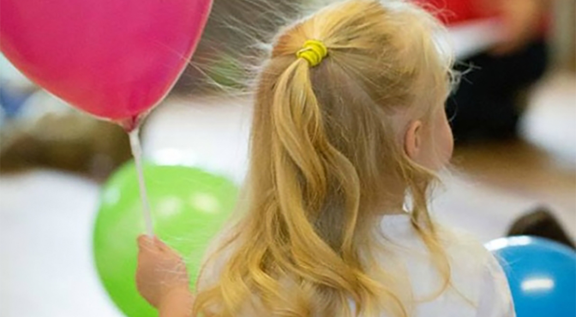 A young girl with blonde ringlets sits holding a pink balloon. She has her back to us and she is sitting looking out at what we assume is a birthday party.